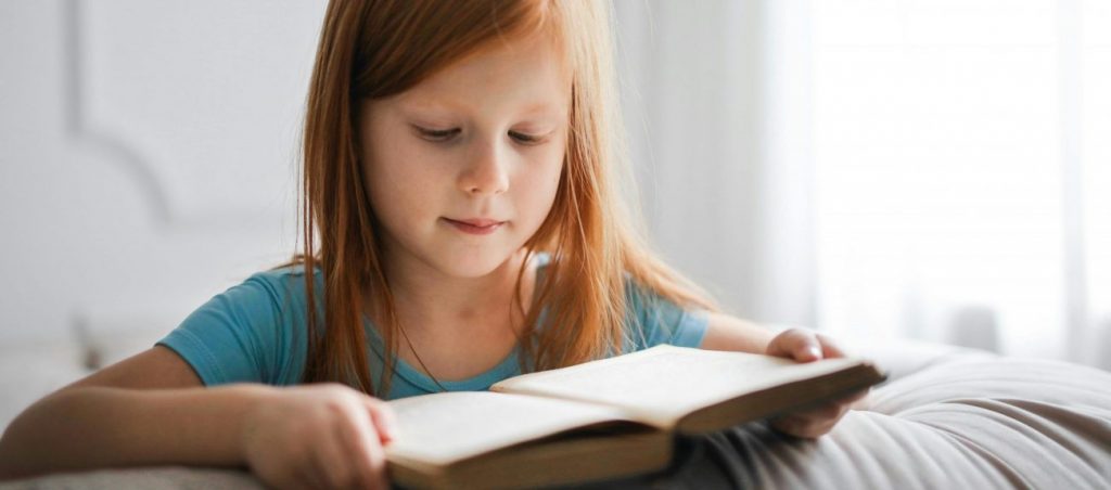 A young girl with red hair is engrossed in reading a book indoors, creating a serene and studious atmosphere.
