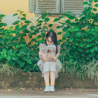 pexels-photo-1196338-1196338 A young girl sits on a bench outdoors, immersed in a book with lush greenery around her.